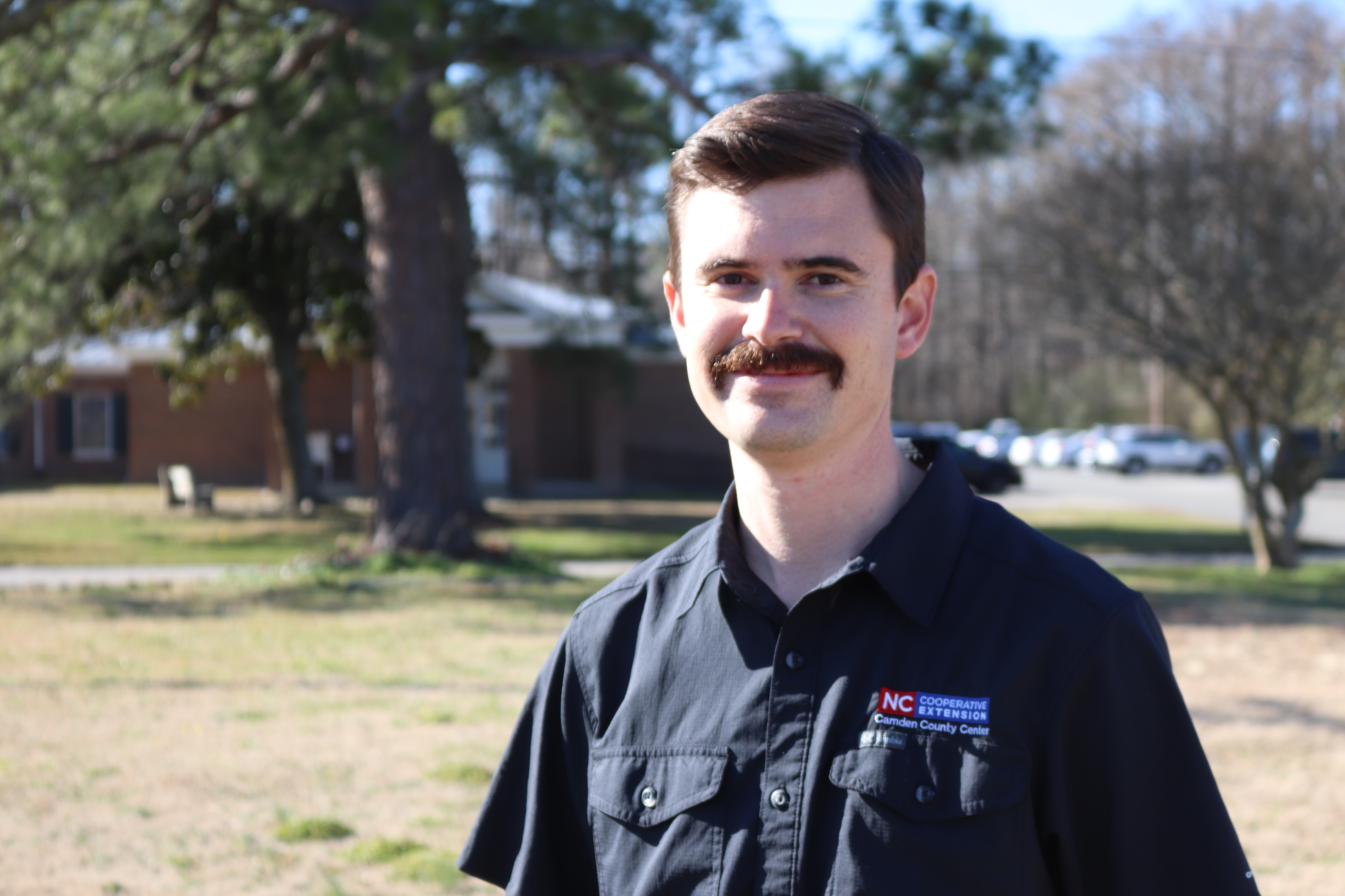 A man in an N.C. Cooperative Extension shirt stands in front of a brick building and trees.